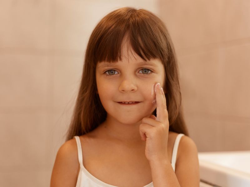 Toma en interiores de una nina con crema facial en la mejilla senalando con el dedo en la cara mirando directamente la camara vistiendo una camiseta blanca sin mangas realizando procedimientos de belleza sola